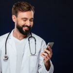 Portrait of a happy male doctor dressed in uniform with stethoscope using mobile phone isolated over dark background