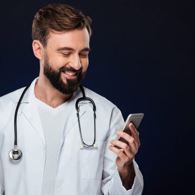 Portrait of a happy male doctor dressed in uniform with stethoscope using mobile phone isolated over dark background